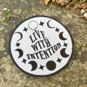 May include: A circular white patch with black embroidered details. The patch features the text "LIVE WITH INTENTION" in a stylized font, surrounded by crescent moons, stars, and a full moon. The patch has a black border.