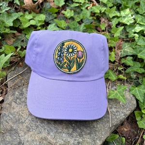 May include: A lavender baseball cap with a floral patch. The patch features a yellow circle with a daisy, tulip, and other flowers. The cap is made of a soft material and has a curved brim.