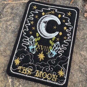 May include: Black rectangular patch featuring a crescent moon, stars, and crystals, embroidered with white, gold, and blue thread. The text "THE MOON" and Roman numeral "XVIII" are also embroidered on the patch. The patch has a white border.