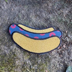 May include: Embroidered patch in the shape of a hot dog. The hot dog bun is yellow, and the hot dog itself is blue with pink circular accents. The patch has a black border.