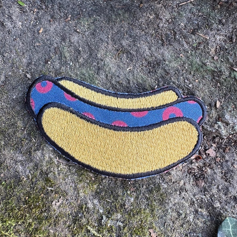 May include: Embroidered patch in the shape of a hot dog. The hot dog bun is yellow, and the hot dog itself is blue with pink circular accents. The patch has a black border.