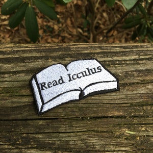 May include: Embroidered patch in the shape of an open book with the words "Read Icculus" in black lettering on a light blue background. The patch has a black border and is set against a weathered wooden surface.