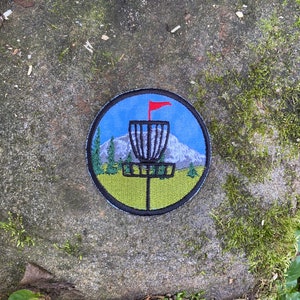May include: Embroidered round patch depicting a disc golf basket with a red flag, a mountain, and trees. The design is set against a blue sky and green grass, with a black border. The patch is suitable for clothing or bags.