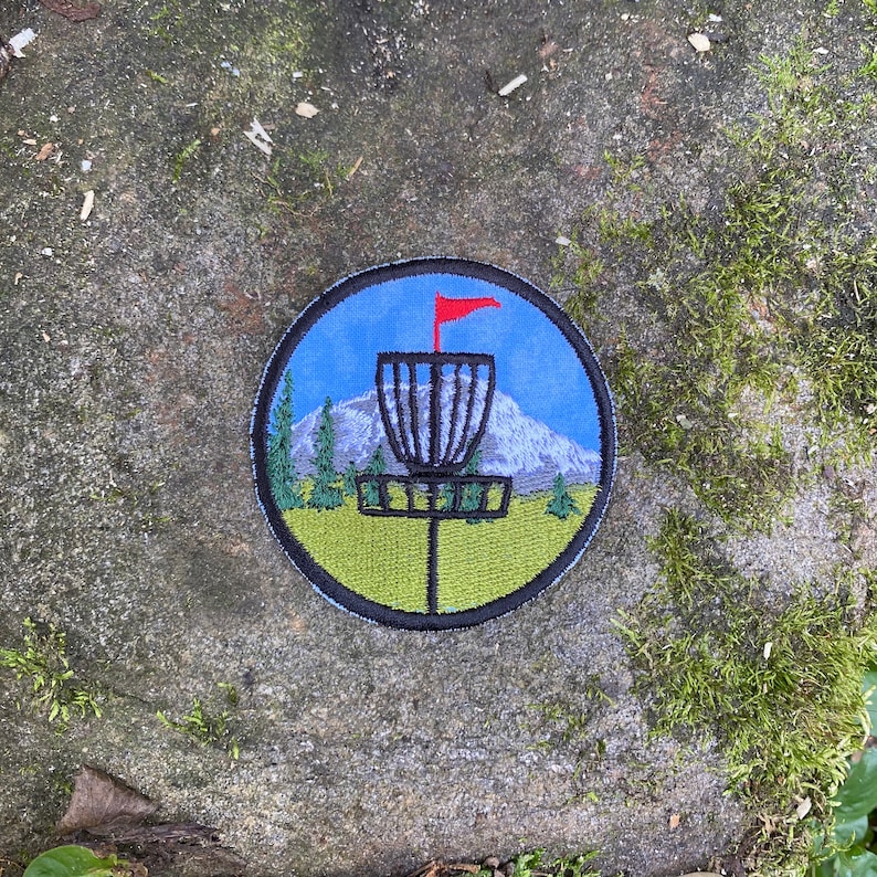 May include: Embroidered round patch depicting a disc golf basket with a red flag, a mountain, and trees. The design is set against a blue sky and green grass, with a black border. The patch is suitable for clothing or bags.