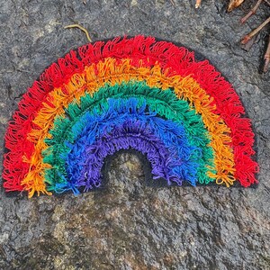 May include: A handmade rainbow-shaped textile art piece. The rainbow features layers of red, orange, yellow, green, blue, and purple fringe. The artwork is set against a dark, textured background, creating a vibrant contrast.