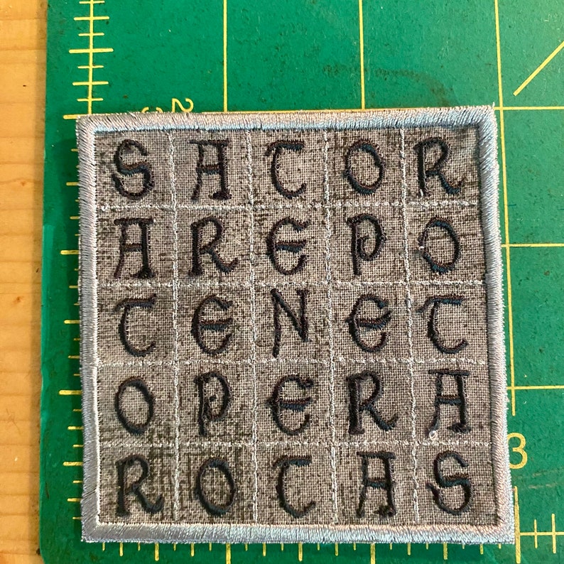 May include: Square embroidered patch with the text "SATOR AREPO TENET OPERA ROTAS" in black letters on a gray background. The patch has a gray border and is on a green surface.