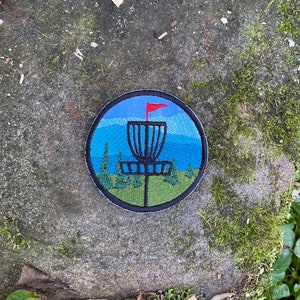 May include: Circular embroidered patch depicting a disc golf basket with a red flag against a blue sky and mountain scene. The patch has a black border and features green trees and grass.