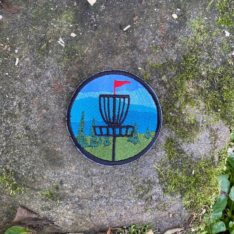 May include: Circular embroidered patch depicting a disc golf basket with a red flag against a blue sky and mountain scene. The patch has a black border and features green trees and grass.