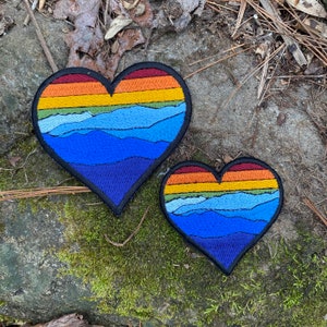 May include: Two heart-shaped patches with a mountain and sunset design. The patches feature a gradient of colours, including blue, orange, yellow, and green. The patches are outlined in black and are placed on a stone surface.