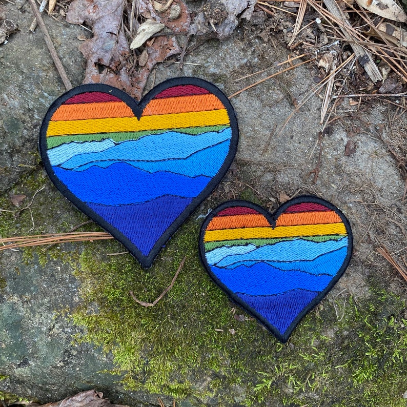 May include: Two heart-shaped patches with a mountain and sunset design. The patches feature a gradient of colours, including blue, orange, yellow, and green. The patches are outlined in black and are placed on a stone surface.