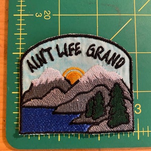 May include: Embroidered patch with the text "AINT LIFE GRAND" above a mountain and lake scene. The patch features a blue sky, grey mountains with white snow, a yellow sun, green trees, and a blue lake. The patch is approximately 7.6 cm wide.