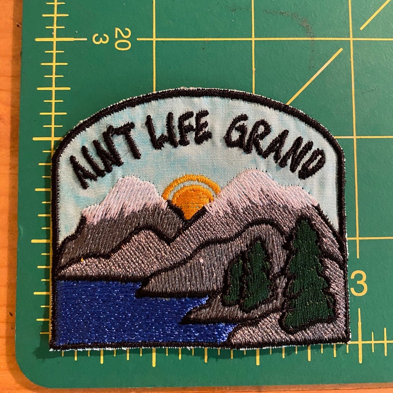 May include: Embroidered patch with the text "AINT LIFE GRAND" above a mountain and lake scene. The patch features a blue sky, grey mountains with white snow, a yellow sun, green trees, and a blue lake. The patch is approximately 7.6 cm wide.