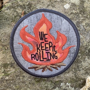 May include: Embroidered patch featuring a stylized campfire with orange and red flames. The text "WE KEEP IT ROLLING" is in the center. The patch has a gray background and a black border, suitable for jackets, bags, or camping gear.