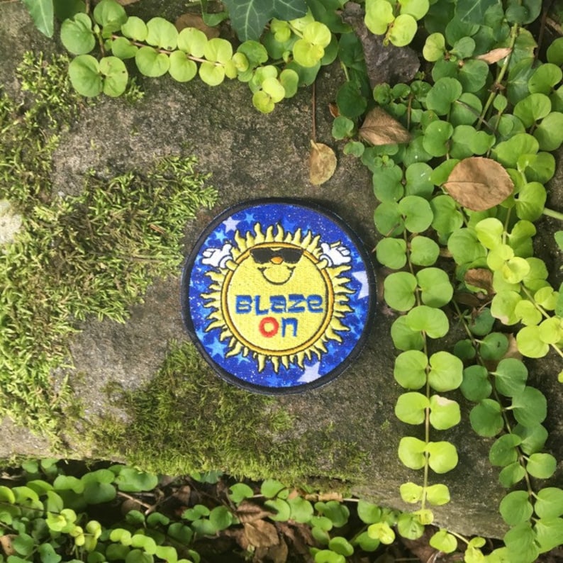 Puede incluir: Un parche redondo con un sol sonriente con gafas de sol, rodeado por un borde azul con estrellas blancas. El texto "BLAZE ON" est&aacute; en el centro. El parche est&aacute; sobre una superficie de piedra con plantas verdes.