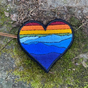 May include: Embroidered heart-shaped patch with a mountain landscape design. The patch features a gradient of colours, including red, orange, yellow, green, and shades of blue. The patch has a black border.