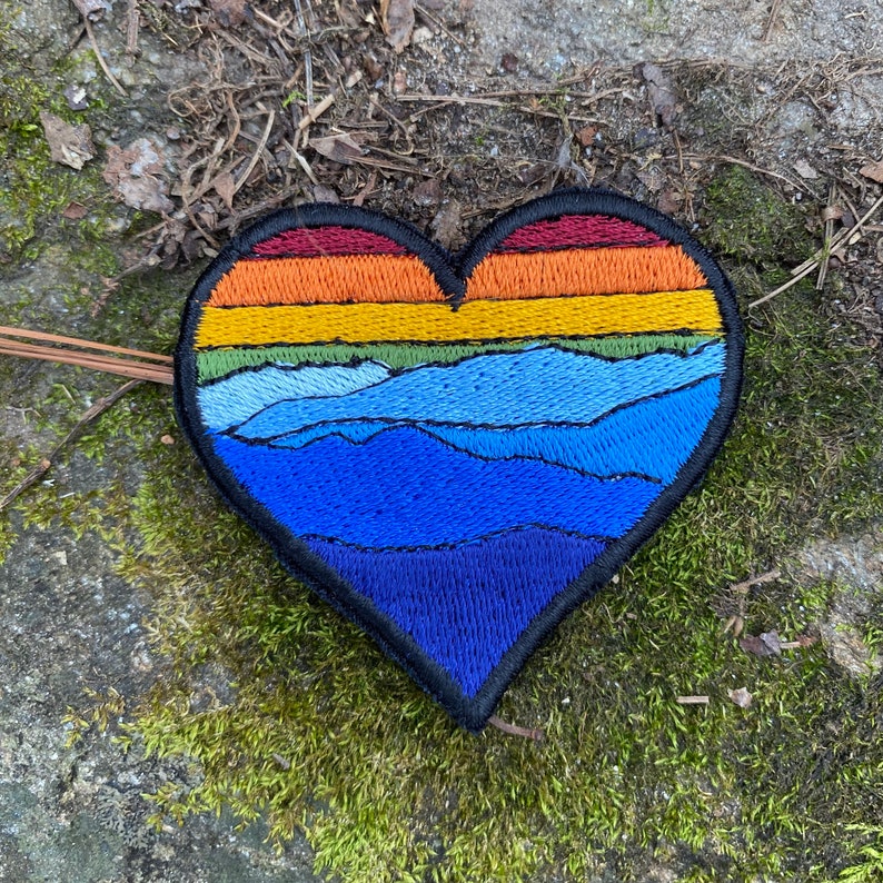 May include: Embroidered heart-shaped patch with a mountain landscape design. The patch features a gradient of colours, including red, orange, yellow, green, and shades of blue. The patch has a black border.