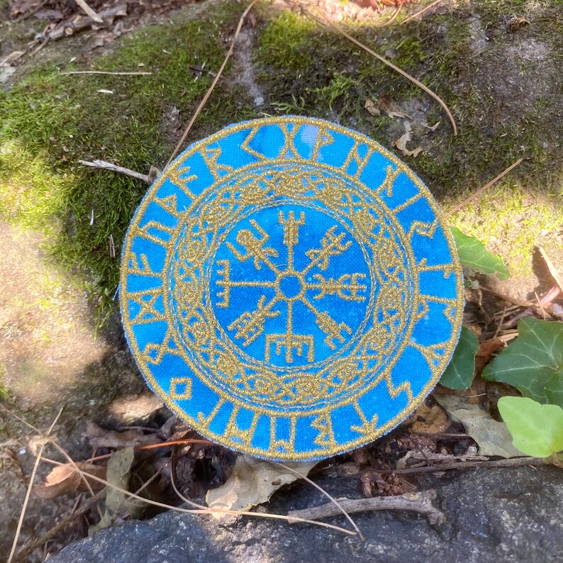 May include: A circular blue patch with gold embroidery. The design features a central symbol surrounded by runic text. The patch is likely made of fabric and is set against a natural background of moss and leaves.