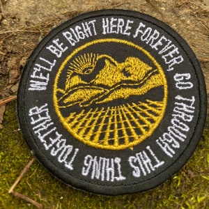 May include: Round black patch with white embroidered text that reads "WE'LL BE RIGHT HERE FOREVER, GO THROUGH THIS THING TOGETHER." The center features a gold embroidered landscape with a sun, mountains, and a grid. The patch is on a natural, mossy surface.