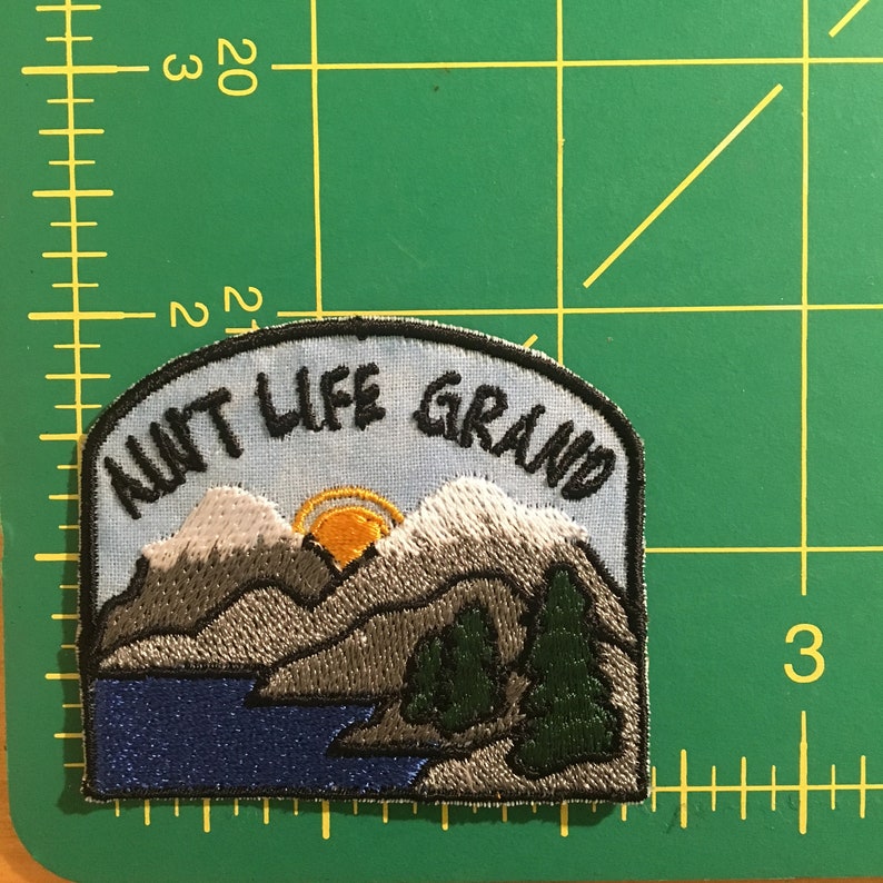 May include: Embroidered patch with a landscape design. The patch features a mountain range, a lake, and pine trees, with a rising sun. The text "AIN'T LIFE GRAND" is arched across the top in black thread. The patch has a light blue background.
