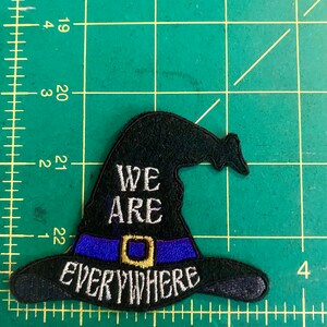May include: Black witch hat patch with the words "WE ARE EVERYWHERE" embroidered in white. The hat has a blue band with a gold buckle. The patch is on a green surface with measurement markings.