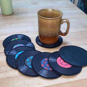 Pu&ograve; includere: Una tazza di ceramica marrone con caff&egrave; poggia su un sottobicchiere nero. Diversi sottobicchieri in vinile nero con etichette centrali colorate sono disposti su una superficie di legno. I sottobicchieri hanno un bordo cucito.