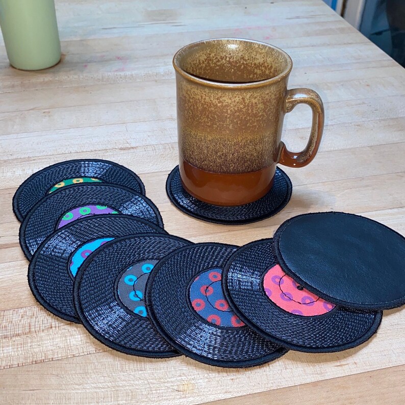 Pu&ograve; includere: Una tazza di ceramica marrone con caff&egrave; poggia su un sottobicchiere nero. Diversi sottobicchieri in vinile nero con etichette centrali colorate sono disposti su una superficie di legno. I sottobicchieri hanno un bordo cucito.