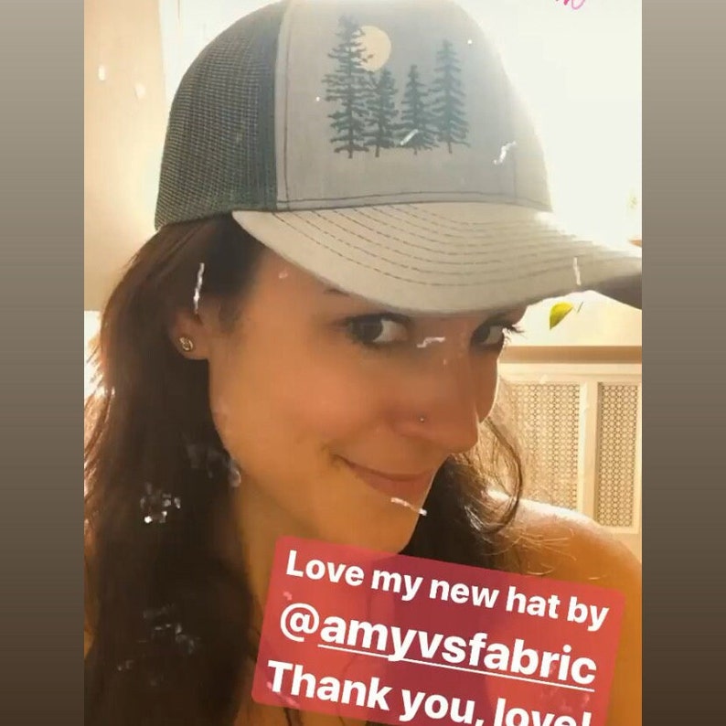 May include: A gray and green trucker hat with a forest and moon design. The hat has a mesh back and a curved brim. The text on the image says "Love my new hat by @amyvsfabric. Thank you, love!"