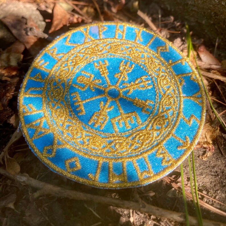 May include: A circular, light blue patch with gold embroidery. The design features a central symbol surrounded by runes and an outer border. The patch is likely made of fabric and is set against a natural background of leaves and twigs.