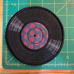 Pu&ograve; includere: Un sottobicchiere circolare nero con una superficie strutturata, che ricorda un disco in vinile. Il centro presenta un tessuto blu con un motivo di cerchi rossi. Cuciture bianche delineano i solchi e i bordi.