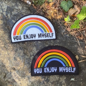May include: Two rainbow-shaped patches with the text "YOU ENJOY MYSELF". One patch has a white background, and the other has a black background. The rainbow design features red, orange, yellow, green, and blue colors.