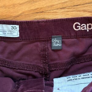 Op de afbeelding: Close-up van de tailleband van een bordeauxrode corduroy broek. Het label luidt "30r, Modern Stretch, True Skinny". Het Gap 1969-logo is zichtbaar. Een klein label luidt "Amy Fabric."