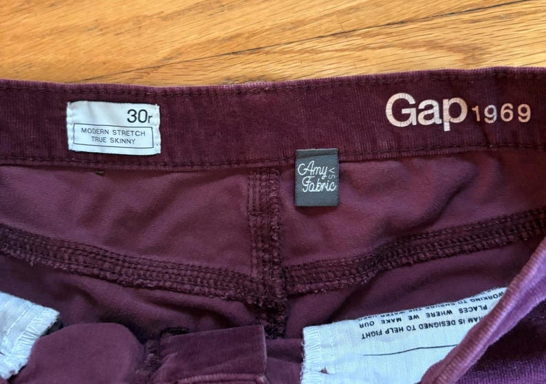 Op de afbeelding: Close-up van de tailleband van een bordeauxrode corduroy broek. Het label luidt "30r, Modern Stretch, True Skinny". Het Gap 1969-logo is zichtbaar. Een klein label luidt "Amy Fabric."