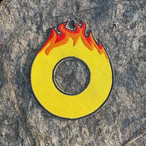 May include: Yellow embroidered patch in the shape of the letter "O" with a black border and flames in red and orange at the top. The patch is circular with a hole in the center.
