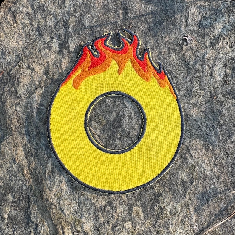 Puede incluir: Parche bordado amarillo con forma de la letra "O" con borde negro y llamas rojas y naranjas en la parte superior. El parche es circular con un agujero en el centro.