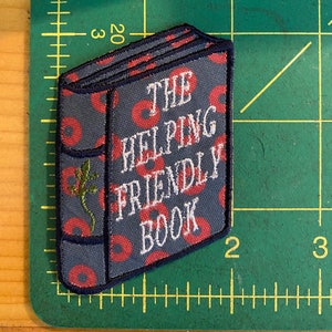 K&ouml;nnte beinhalten: Gestickter Aufn&auml;her in Buchform mit dem Text "THE HELPING FRIENDLY BOOK". Der Aufn&auml;her hat einen grauen Hintergrund mit roten Kreisen und einem gr&uuml;nen Eidechsen-Motiv. Die R&auml;nder sind marineblau.