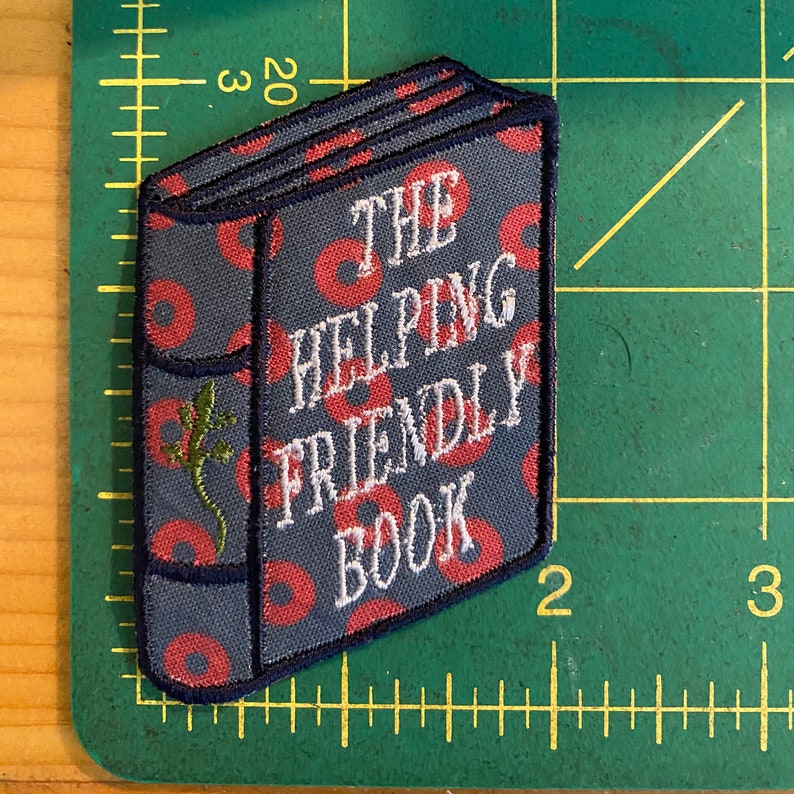K&ouml;nnte beinhalten: Gestickter Aufn&auml;her in Buchform mit dem Text "THE HELPING FRIENDLY BOOK". Der Aufn&auml;her hat einen grauen Hintergrund mit roten Kreisen und einem gr&uuml;nen Eidechsen-Motiv. Die R&auml;nder sind marineblau.