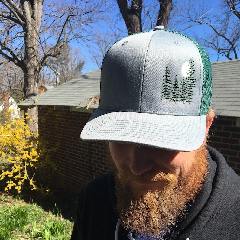 May include: A gray trucker hat with a green mesh back. The front panel features an embroidered design of pine trees and a full moon. The hat has a curved brim and a snapback closure.
