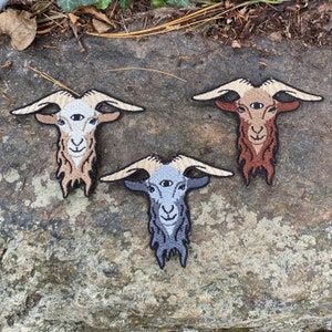May include: Three embroidered patches featuring a goat head with a third eye. The patches are in shades of tan, brown, and gray, with cream-colored horns. The goat's face has a smiling expression. The patches are on a stone surface.