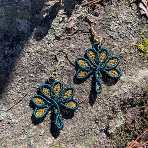 Pode incluir: Um par de brincos feitos &agrave; m&atilde;o com um design de trevo de tr&ecirc;s folhas. Os brincos t&ecirc;m um contorno verde-azulado escuro com filigrana dourada. Os ganchos para as orelhas s&atilde;o prateados.