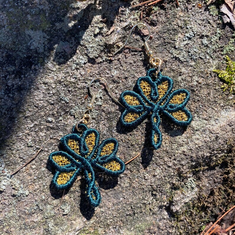 Pode incluir: Um par de brincos feitos &agrave; m&atilde;o com um design de trevo de tr&ecirc;s folhas. Os brincos t&ecirc;m um contorno verde-azulado escuro com filigrana dourada. Os ganchos para as orelhas s&atilde;o prateados.