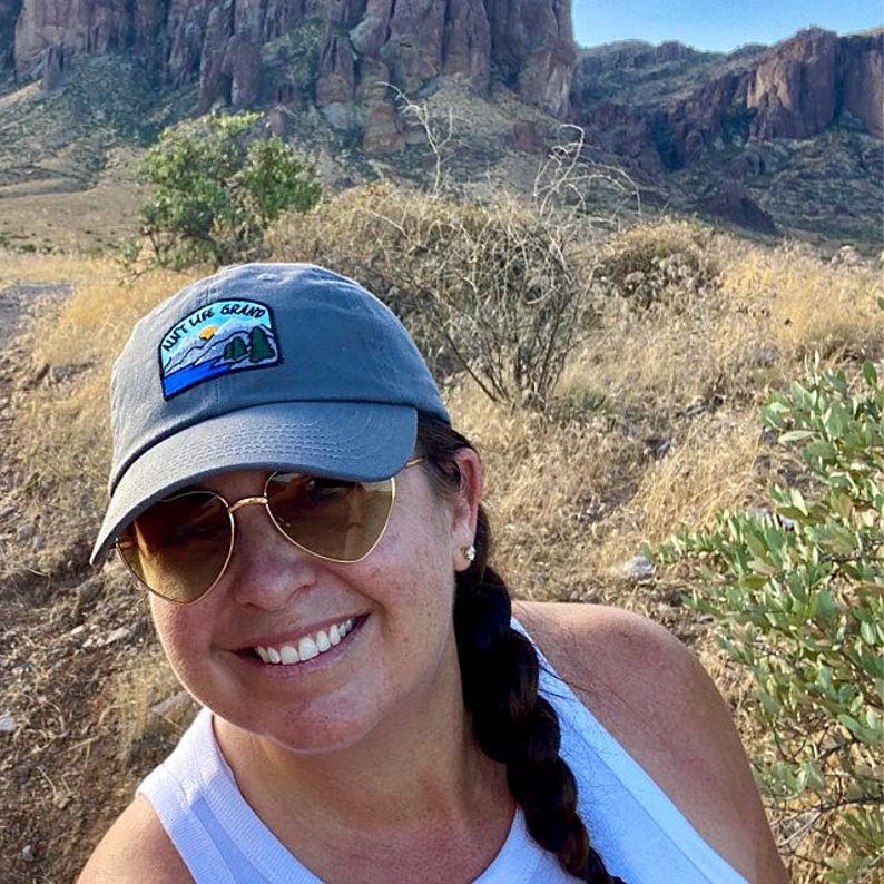 May include: Grey baseball cap with a patch reading "Ain't Life Grand", featuring a mountain and tree design. The cap is worn with heart-shaped sunglasses.