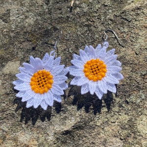 May include: A pair of daisy-shaped earrings. Each earring features a white, lace-like petal design surrounding a yellow center. The earrings have silver-colored hooks for pierced ears. The earrings are displayed on a textured, gray surface.