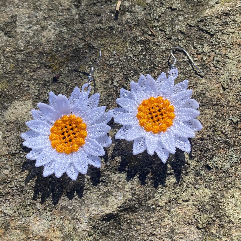 May include: A pair of daisy-shaped earrings. Each earring features a white, lace-like petal design surrounding a yellow center. The earrings have silver-colored hooks for pierced ears. The earrings are displayed on a textured, gray surface.