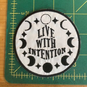 May include: Circular white patch with a black embroidered design. The patch reads "LIVE WITH INTENTION" in a decorative font, encircled by crescent moons, stars, and a full moon. It has a black border and is on a green surface.