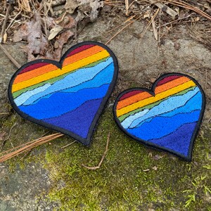 May include: Two heart-shaped embroidered patches. The patches feature a mountain range design with a gradient of colours, including red, orange, yellow, and blue. The patches are outlined in black and are placed on a mossy surface.