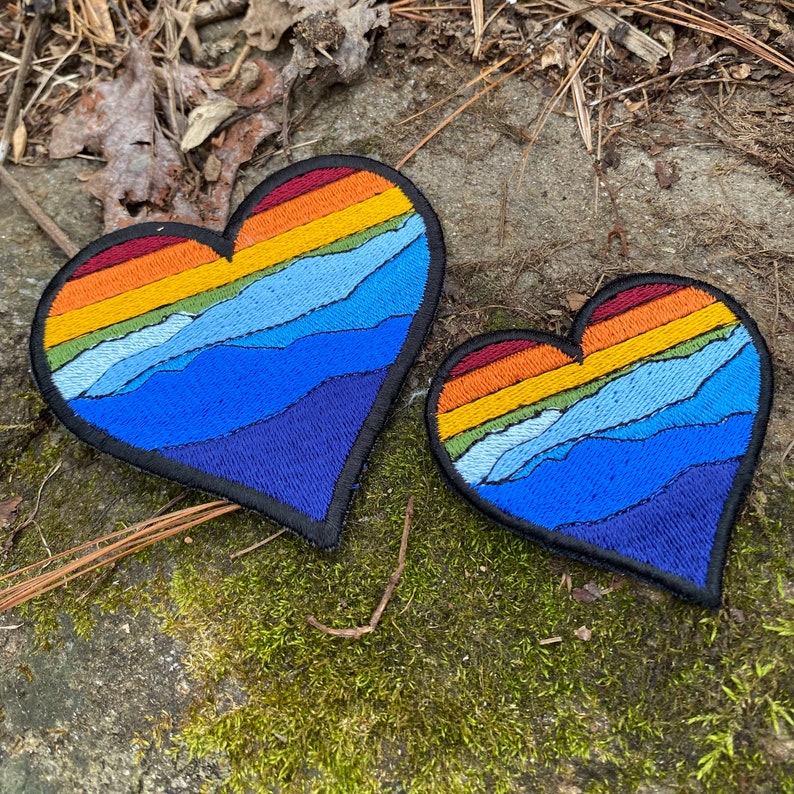 May include: Two heart-shaped embroidered patches. The patches feature a mountain range design with a gradient of colours, including red, orange, yellow, and blue. The patches are outlined in black and are placed on a mossy surface.
