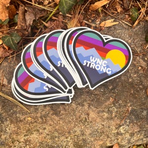 Puede incluir: Pegatinas en forma de coraz&oacute;n con un dise&ntilde;o de monta&ntilde;a y puesta de sol. Las pegatinas presentan una paleta de colores p&uacute;rpura, rojo, azul y amarillo. El texto "WNC STRONG" est&aacute; impreso en blanco. Las pegatinas est&aacute;n apiladas sobre una superficie de piedra.