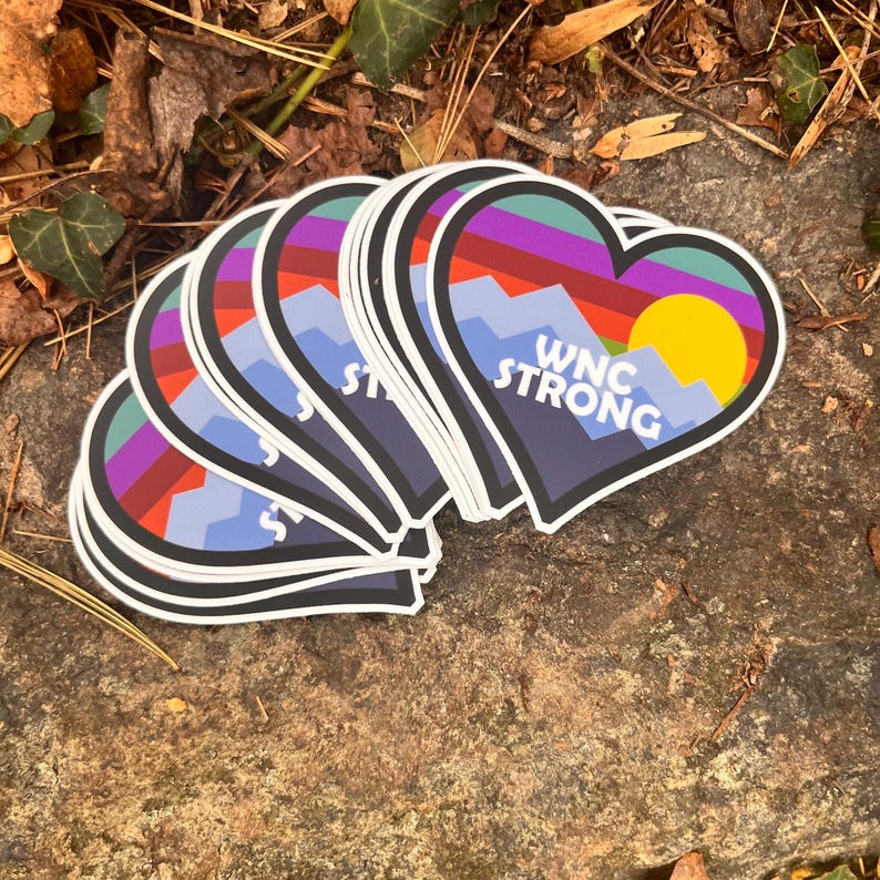Puede incluir: Pegatinas en forma de coraz&oacute;n con un dise&ntilde;o de monta&ntilde;a y puesta de sol. Las pegatinas presentan una paleta de colores p&uacute;rpura, rojo, azul y amarillo. El texto "WNC STRONG" est&aacute; impreso en blanco. Las pegatinas est&aacute;n apiladas sobre una superficie de piedra.