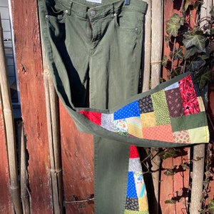 May include: Olive green denim jeans with a patchwork design on one leg. The patchwork features various fabric patterns and colors, including red, blue, and floral prints. The jeans are hanging on a clear hanger.