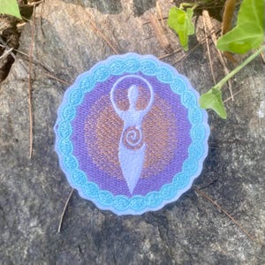 May include: Embroidered patch with a white figure in a purple and gold circular design, bordered by a light blue scalloped edge. The patch is set on a grey stone surface with green foliage. The design depicts a stylized female form.